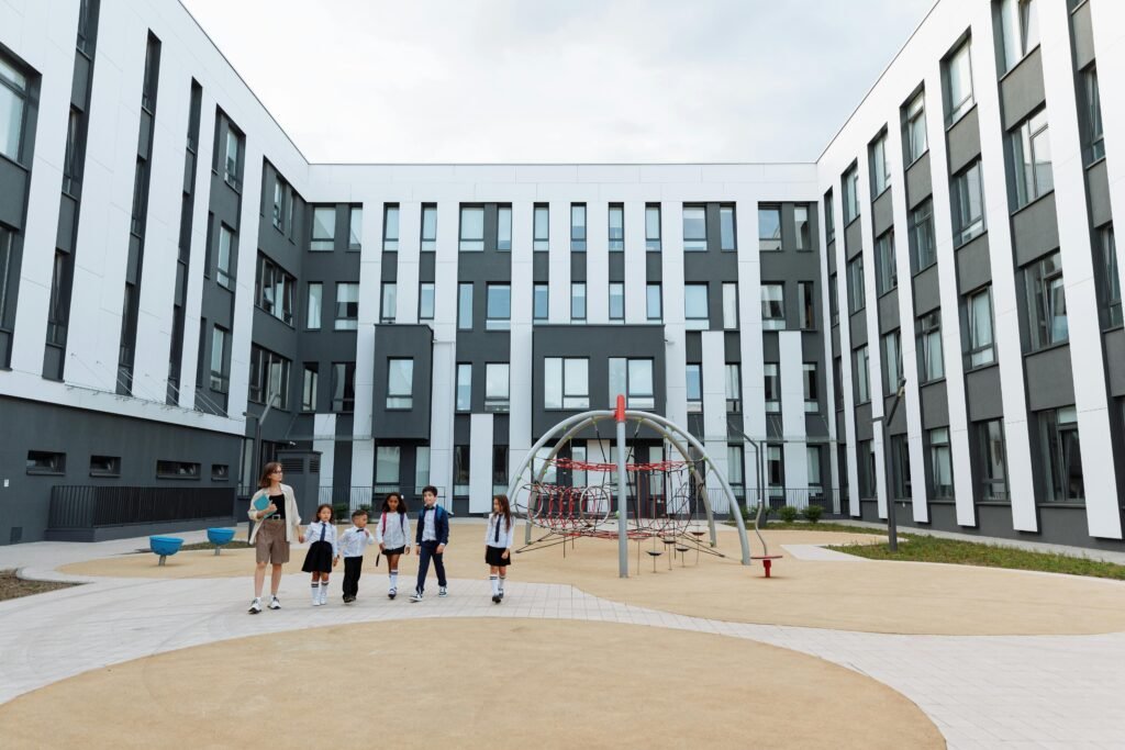 Group of students and teacher walking in a school courtyard with modern architecture and playground.